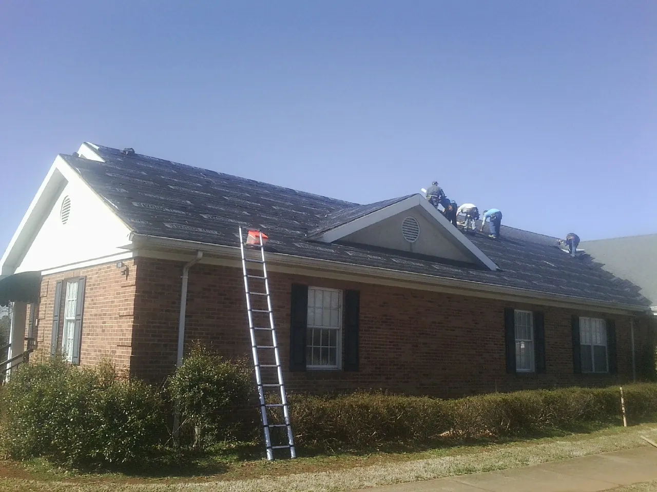 Crew tearing shingles off a brick ranch roof with a ladder set — Metal Roof Repair work by our crew in Annabella