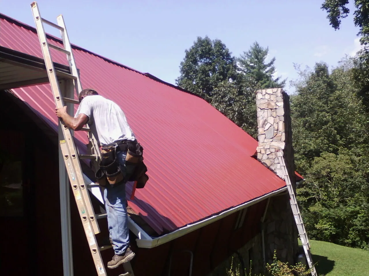 Roofer on a ladder beside a red metal roof and stone chimney — Soffit & Fascia Repair by Roofing Brothers in Annabella